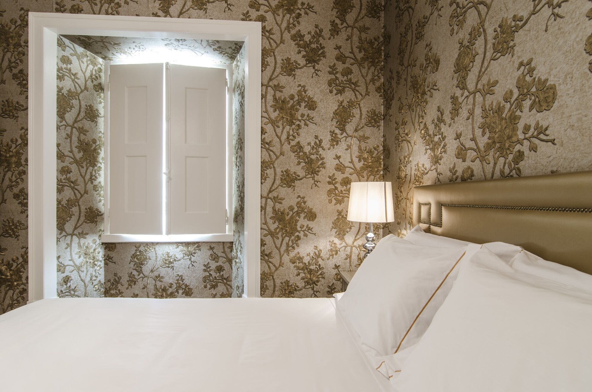 Et hotelværelse med en seng, et natbord, en lampe og en dør med blomstret tapet.