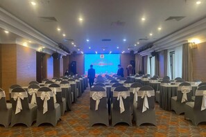 Un salón de conferencias con filas de mesas y sillas, un escenario con una pantalla para presentaciones y suelo alfombrado.