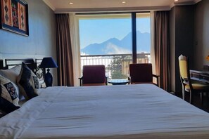 Habitación de hotel con una cama grande, dos sillas, una mesita y vistas a montañas desde la ventana.