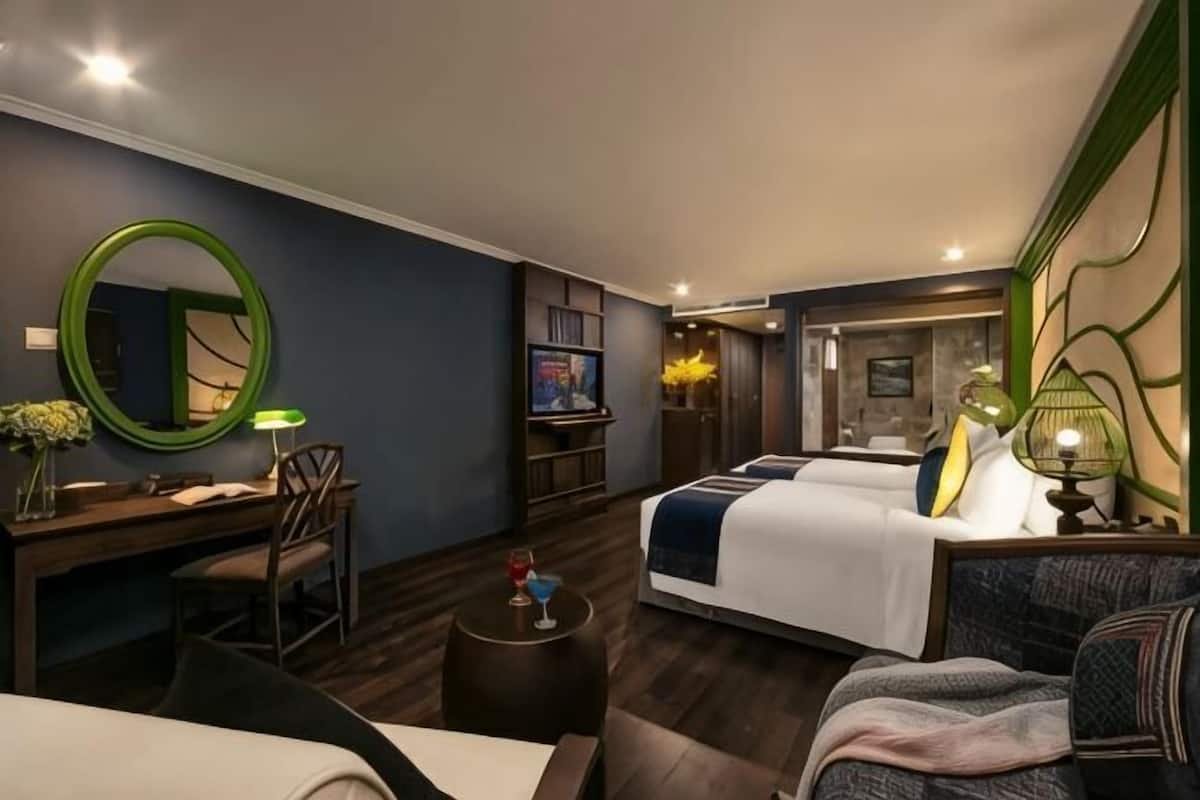 A hotel room with a bed, a desk, a chair, a round mirror, and a small table with a lamp.