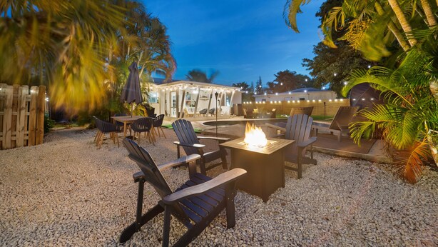 Een terras met Adirondack-stoelen en een vuurkuil, omgeven door palmbomen en een grindpad.