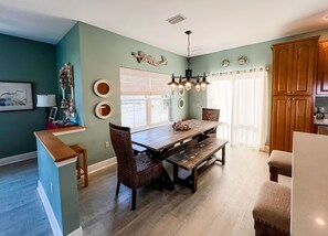 Uma sala de jantar com mesa e cadeiras de madeira, um candelabro e vista para o exterior através de grandes janelas.