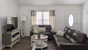 A living room with a dark leather sofa, a white coffee table, a television on a stand, and a door with a frosted glass window. - Lake Life Getaway! (Winchester)