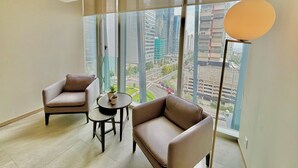 Una habitación moderna con dos sillones de cuero, una mesita redonda y una lámpara de pie. Amplias ventanas ofrecen vistas a un paisaje urbano con edificios altos y una carretera.