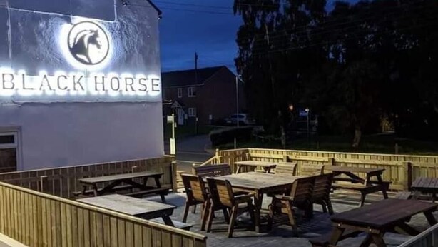 An outdoor seating area with wooden tables and chairs, a neon sign for 'BLACK HORSE', and a wooden fence. - The Stables at The Black Horse (Crook)