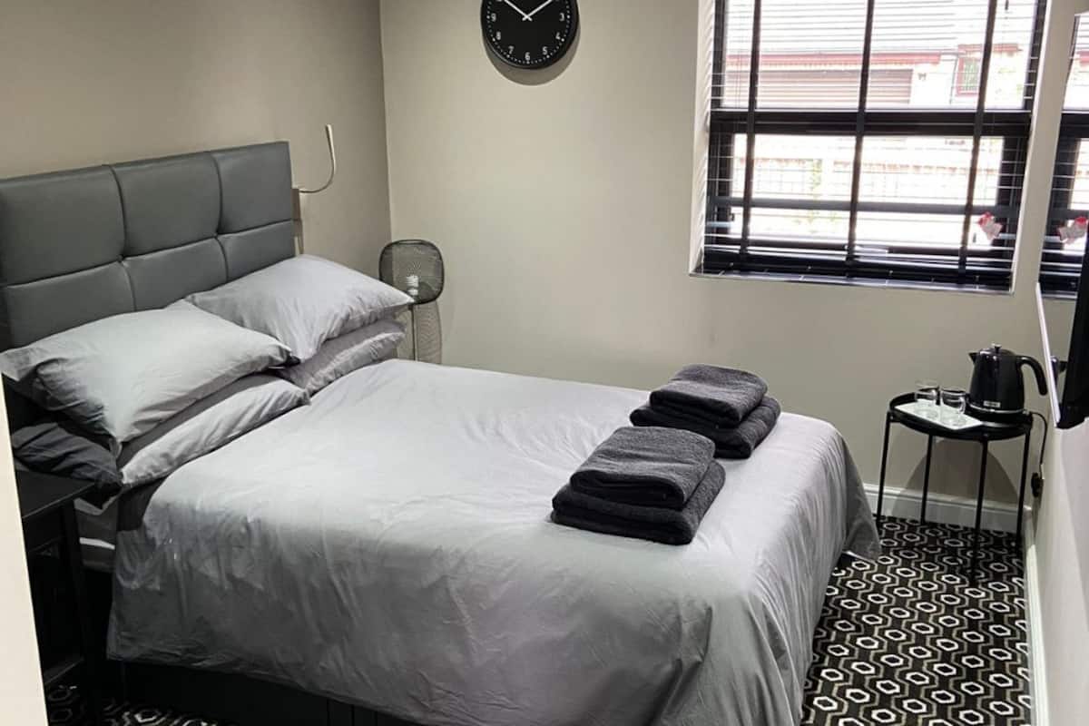 Una habitación de hotel moderna con una cama grande, un reloj de pared, una ventana con persianas y una pequeña mesa con hervidor.