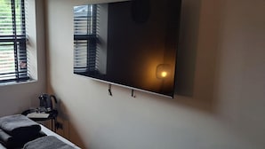 A hotel room with a bed, a flat-screen TV mounted on the wall, a small table with a kettle and glasses, and a window with blinds. - The Stables at The Black Horse (Crook)