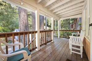 A covered porch with wooden railings, white chairs, and a view of a forested area.