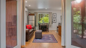 Un salon chaleureux avec un canapé en cuir marron, un petit pouf et une table à manger avec des chaises. On aperçoit une fenêtre avec des rideaux, et un réfrigérateur se trouve au fond de la pièce.