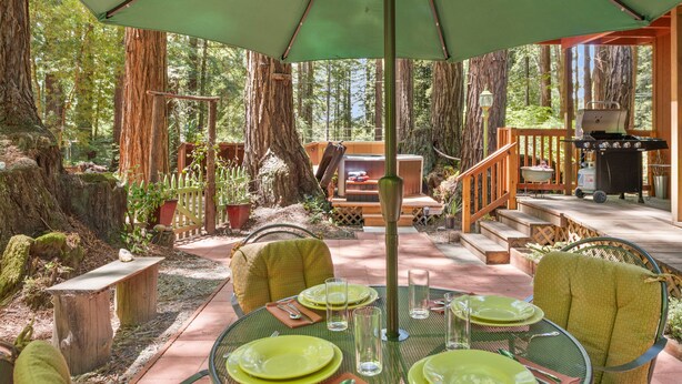 Un espace extérieur aménagé pour les repas, avec une table dressée pour quatre personnes, entouré de grands arbres et doté d’une terrasse en bois.