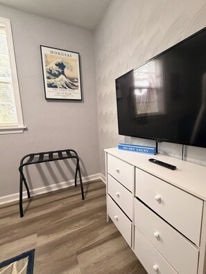 Une pièce dotée d’un grand téléviseur à écran plat fixé au mur, d’une commode blanche à tiroirs, d’un banc en métal noir et d’une œuvre d’art encadrée intitulée « Hokusai : la Grande Vague » qui orne le mur.