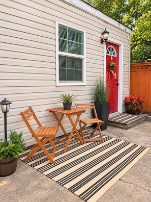 A small house with a red door, a window with blinds, a wooden table and chairs, a black lamp, and a striped rug.