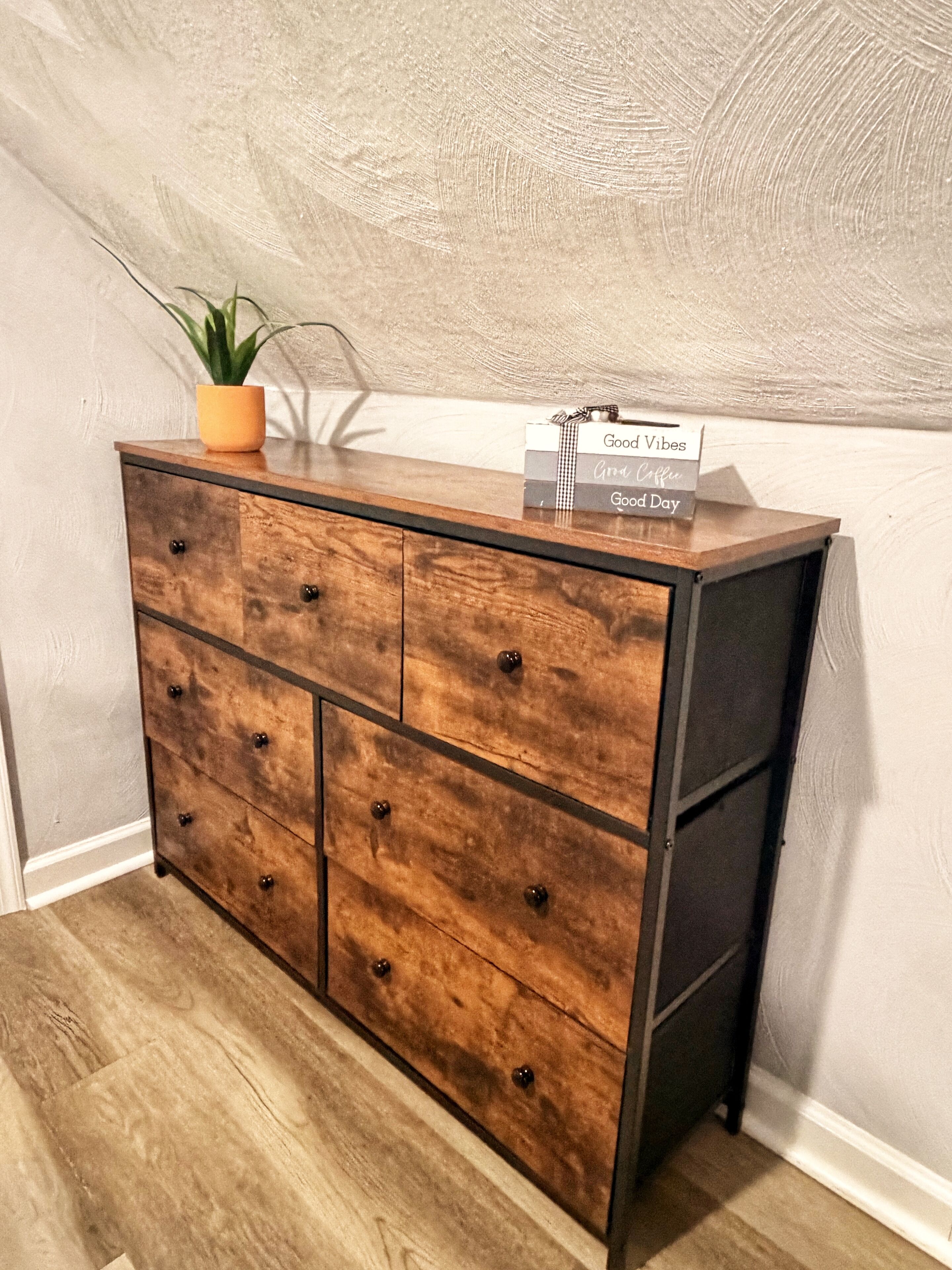 A wooden dresser with drawers and knobs, a potted plant, and a sign with positive messages.