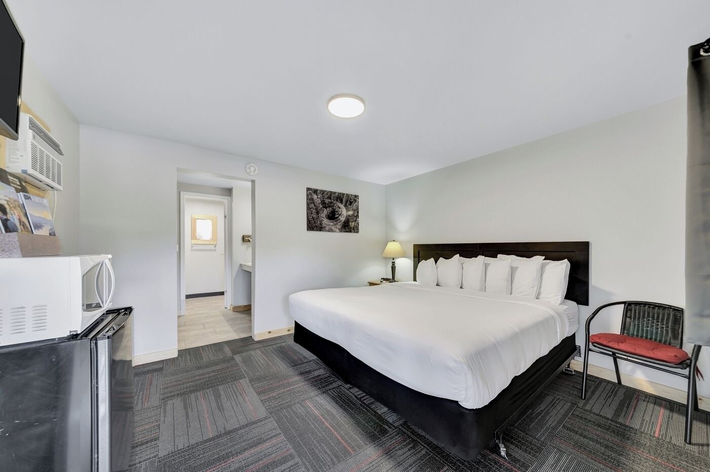 Une chambre d’hôtel avec un grand lit, une chaise, un bureau avec une lampe, un réfrigérateur et une porte donnant sur une autre pièce.
