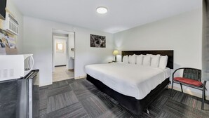 A hotel room with a large bed, a chair, a desk with a lamp, a refrigerator, and a door leading to another room.