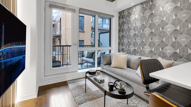 Una sala de estar moderna con un sofá, una mesa de centro y un televisor. Hay ventanas amplias con vistas a edificios y un balcón.