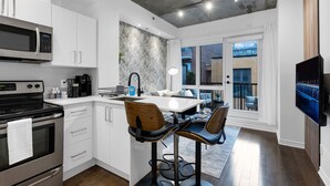 A modern kitchen with white cabinetry, a built-in microwave, and a stainless steel stove. There is a dining area with a white table and two bar stools. - Suite 407 Downtown Montreal | W/ Balcony, Queen Bed, Sofa Bed, Parking & 55` TV (Montréal)