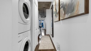 A laundry room with two front-loading washers and dryers, a wooden floor, and a hallway leading to a room with a blue window and a chair. - Suite 407 Downtown Montreal | W/ Balcony, Queen Bed, Sofa Bed, Parking & 55` TV (Montréal)
