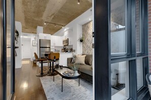 A modern kitchen with white cabinets, a microwave, and a coffee machine. There is a dining area with a table and chairs, and a sofa in the living room. - Suite 407 Downtown Montreal | W/ Balcony, Queen Bed, Sofa Bed, Parking & 55` TV (Montréal)