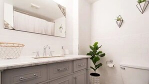 Un baño con lavamanos, espejo y retrete blancos. Hay dos plantas en macetas y un taburete pequeño.