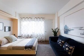 Una camera d'albergo con un letto, un divano blu scuro, una finestra con tende a motivi e un quadro appeso al muro.