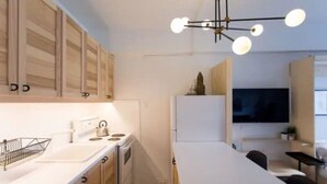 Una cocina moderna con armarios de madera, encimera blanca, un hervidor y un televisor al fondo.