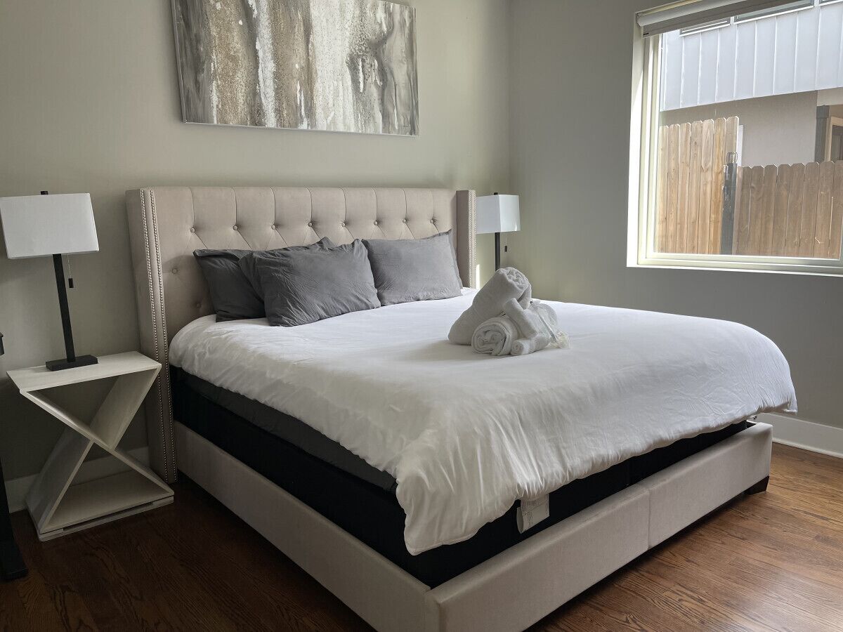 A neatly made bed with a white headboard and grey bedding, a bedside table with a lamp, and a window with a view of a wooden fence.
