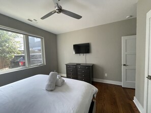 A bedroom with a bed, a dresser, a ceiling fan, a TV mounted on the wall, and a door.