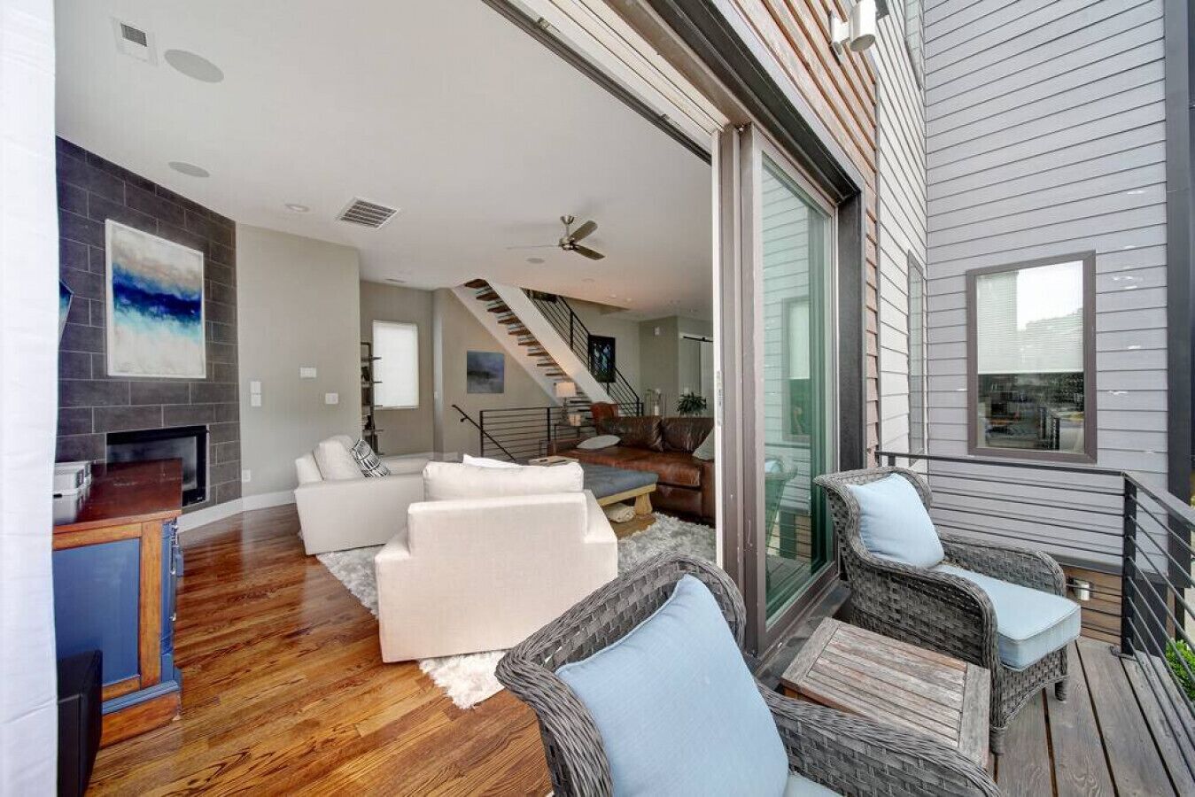 A modern living room with a fireplace, white sofas, wicker chairs, and a staircase leading to an upper level.
