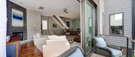 A modern living room with a fireplace, white sofas, wicker chairs, and a staircase leading to an upper level.