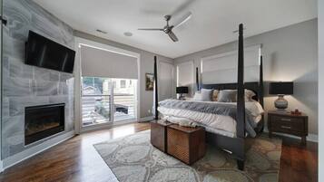 A bedroom with a fireplace, a bed, a nightstand, a ceiling fan, and a large window.