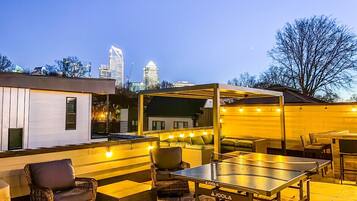 An outdoor patio with seating and a ping pong table, surrounded by a white wall and a city skyline in the background.