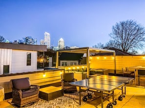 En udendørs terrasse med siddepladser og et pingpongbord, omgivet af en hvid mur og en bys skyline i baggrunden.