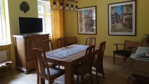 Uma sala de jantar com uma mesa de madeira, cadeiras, uma televisão num armário e duas molduras na parede.