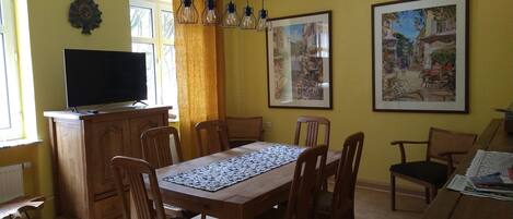 A dining area with a wooden table, chairs, a television on a cabinet, and two framed pictures on the wall.