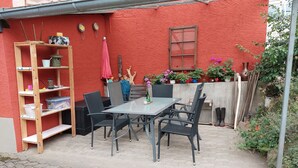 Um pátio com uma mesa e cadeiras, uma prateleira de madeira com diversos objetos e uma parede vermelha com uma janela e plantas em vasos.