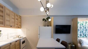 A compact kitchen with wooden cabinets, a white countertop, and a built-in oven. There is a dining table with chairs, a TV mounted on the wall, and a bed with a patterned curtain.