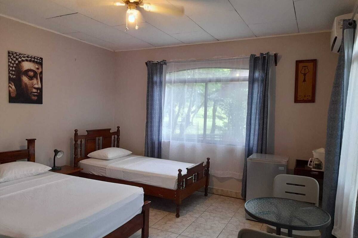 A room with two wooden beds, a small table, a chair, a ceiling fan, and a window with curtains.