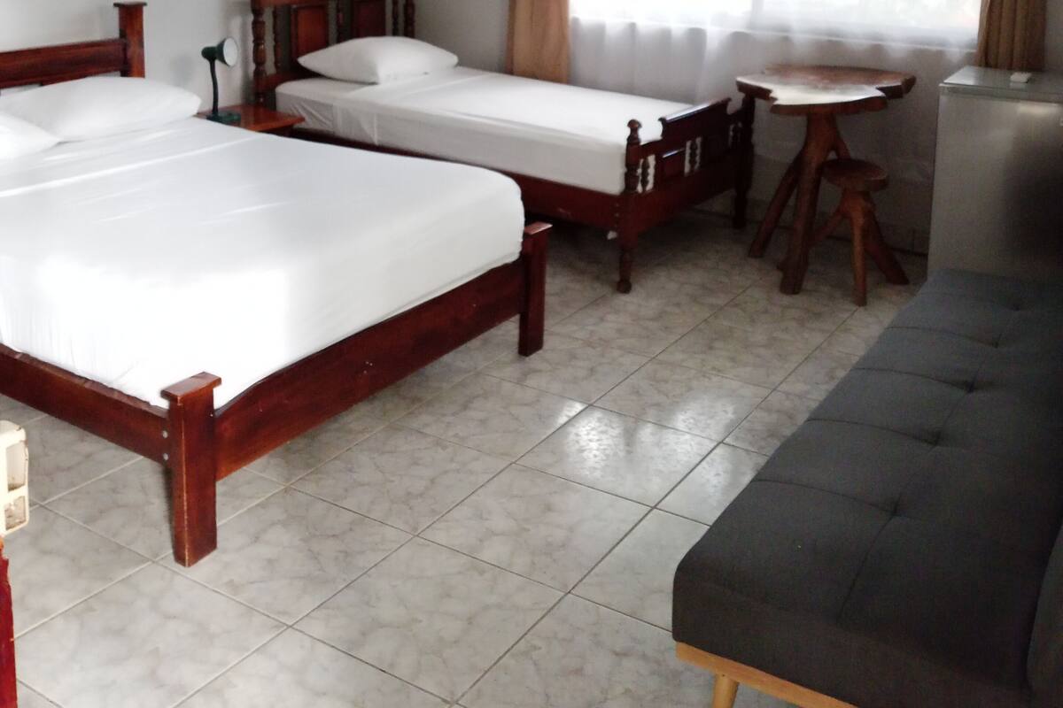 A hotel room with two wooden beds, a grey sofa, a small table, and a ceiling fan.