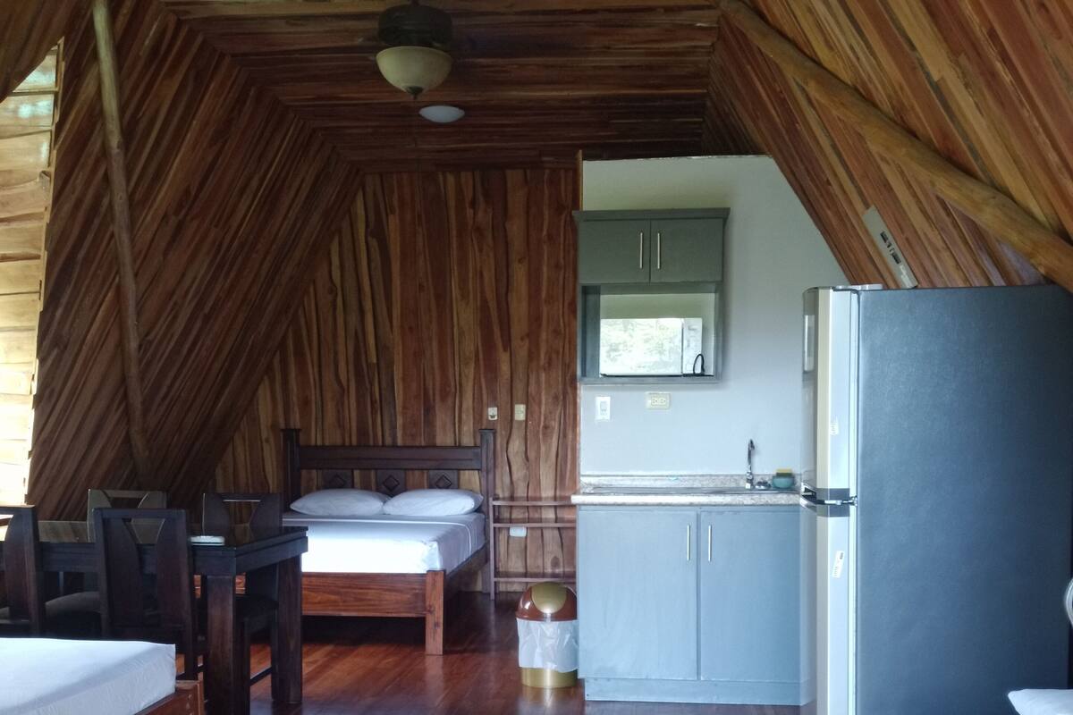 A room with a sloped ceiling, wooden walls, a bed, a desk, a chair, a small table, a refrigerator, and a window.