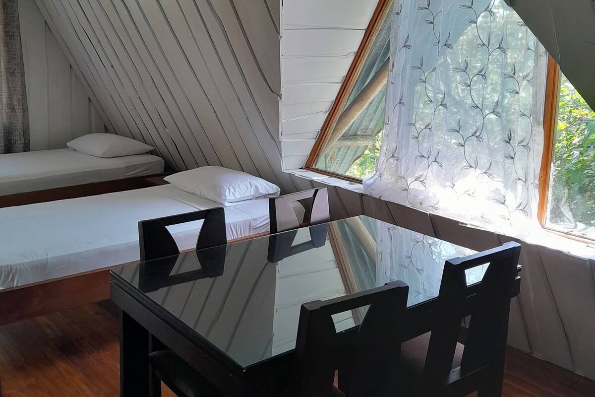 A room with a sloped ceiling, a bed with white bedding, a dining table with chairs, and a window with a patterned curtain.