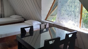 A room with a sloped ceiling, a bed with white bedding, a dining table with chairs, and a window with a patterned curtain.
