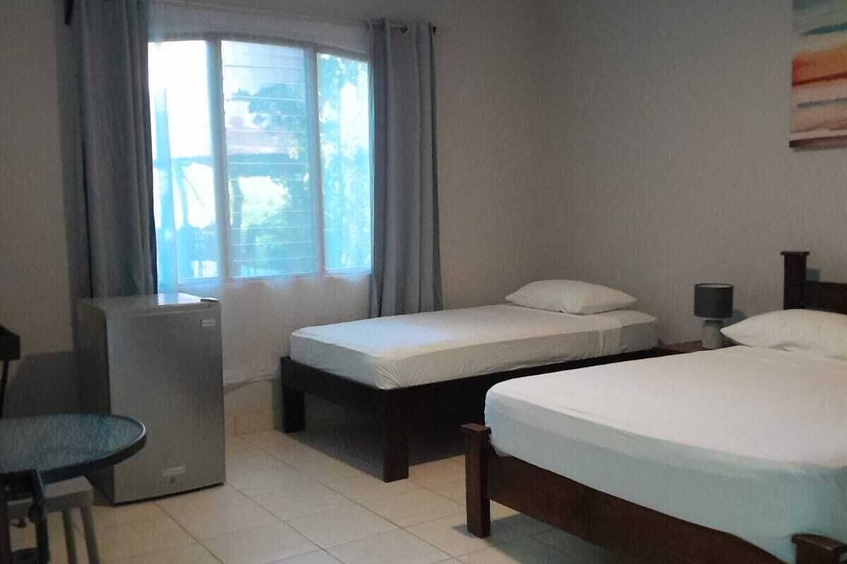 A hotel room with two single beds, a larger bed, a desk with a chair, a small table, and a window with curtains.