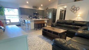 A modern living room with a black leather sofa, a wooden coffee table, and a patterned rug. There is a kitchen area with grey cabinets and a bar counter.