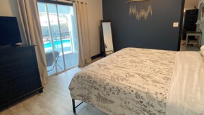 A bedroom with a large bed, a ceiling fan, a view of a swimming pool, and a dark wall with a decorative hanging.