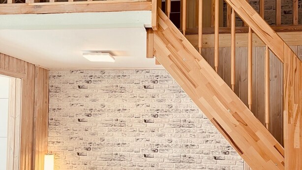 A modern loft with exposed wooden beams, a brick-patterned wall, a wooden staircase, a grey sectional sofa, and a glass coffee table. - Extertal-Ferienpark - PREMIUM HOLIDAY HOUSE - Sauna, fireplace, hiking, relaxation #49 (Extertal)