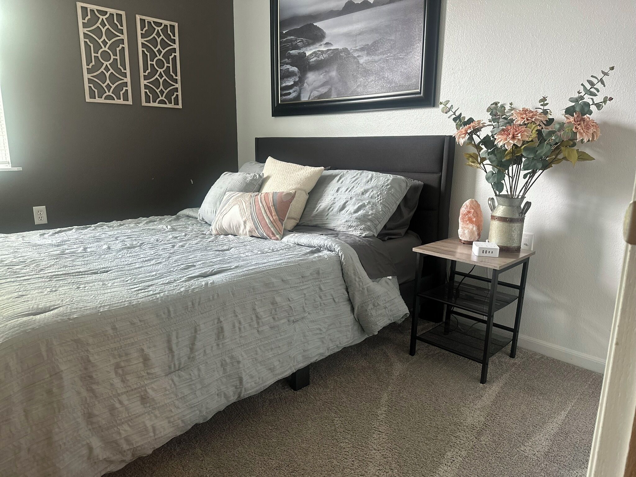 Un letto rifatto con un copriletto grigio e numerosi cuscini, un comodino con una lampada di sale e un vaso di fiori, e una fotografia incorniciata appesa al muro.