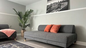 Un salon moderne avec un canapé gris, une grande plante en pot et un tableau représentant un paysage urbain en noir et blanc accroché au mur.