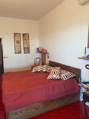 A bedroom with a wooden bed frame, a red bedspread, and patterned pillows. There are two wall-mounted shelves with books and decorative items.