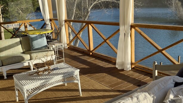 Terraza de madera con vista a un lago, ventilador de techo y una zona de estar con sofá y mesa de mimbre.